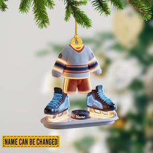 May include: A hockey-themed Christmas ornament featuring a miniature jersey and shorts, ice skates, and a hockey puck. The jersey is white with blue and orange stripes. The skates have blue laces. The puck has a name on it. The text "NAME CAN BE CHANGED" is at the bottom.