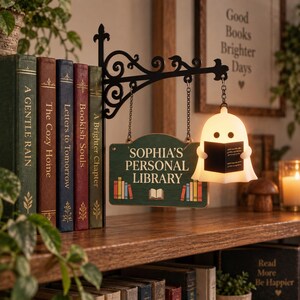May include: A black metal wall-mounted bracket holds a green rectangular sign that reads SOPHIA'S PERSONAL LIBRARY with a book graphic. A white glowing ghost-shaped lamp hangs from the bracket, holding a small black book in its hands.