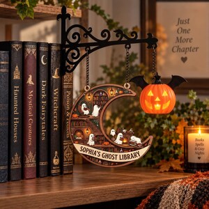 May include: A decorative wall-mounted sign featuring a crescent moon shape with a library scene filled with small white ghosts, hanging from a black metal bracket. An orange jack-o'-lantern with black bat wings hangs beside it. Text reads: SOPHIA'S GHOST LIBRARY.