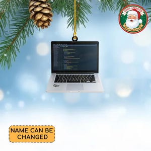 May include: A silver laptop Christmas ornament with a screen displaying code. The ornament hangs from a gold chain, with a Santa Approved seal in the background. The text "NAME CAN BE CHANGED" is on a yellow rectangle.