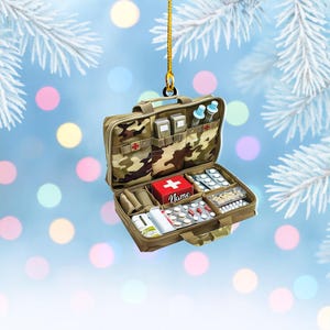 May include: A camouflage-patterned first aid kit Christmas ornament. The open kit reveals various medical supplies, including pills, bandages, and vials. A red box with a white cross and the name "Name" is visible. The ornament hangs from a gold string.