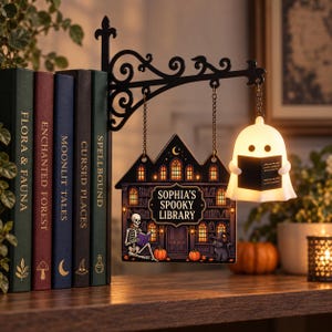 May include: A black metal wall bracket holds a hanging sign that reads "SOPHIA'S SPOOKY LIBRARY" with a skeleton and cat illustration. Beside it, a small white glowing ghost figure holds a book. The sign hangs next to five decorative books.