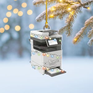 May include: A white and gray office copier Christmas ornament with a gold hanging loop. The ornament is decorated with a string of colorful lights and has the word "Name" printed on the front.