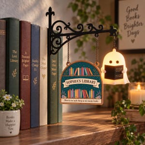 May include: A black metal wall bracket holds a hanging wooden sign labeled SOPHIA'S LIBRARY with colorful book illustrations and the text There is no such thing as too many books. A small, glowing white ghost figure holds a black book next to the sign.