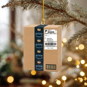 May include: A brown cardboard box ornament with a blue "prime" stripe and logo. The ornament has a shipping label that reads "Priority Mail 1-Day" and "Happy Birthday, JU". The ornament is hanging from a gold string.
