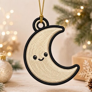 May include: A crescent moon ornament featuring cream-colored embroidery with a black stitched border and a smiling face with two black eyes. The ornament is suspended by a thin gold cord attached to a circular loop at the top of the moon shape.