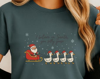 May include: Dark teal t-shirt with a Christmas graphic. The design features Santa Claus in a sleigh pulled by geese wearing Santa hats, with the text "I believe in Santa, you silly goose."