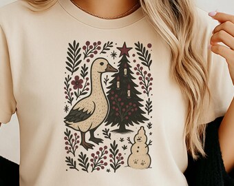 May include: Beige t-shirt featuring a whimsical illustration of a goose, Christmas tree, and snowman. The design is framed by floral elements in burgundy and green. The shirt has short sleeves and a crew neck.