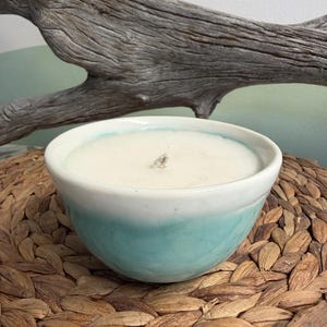 May include: A white wax candle in a ceramic bowl with a smooth white interior and a gradient turquoise blue exterior. The bowl has a rounded shape and a single centred wick. The candle is filled to the brim with solid white wax.
