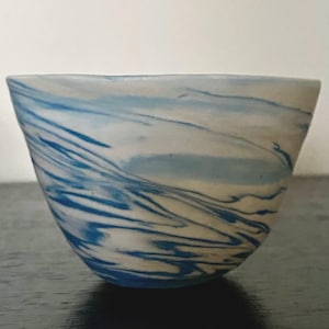 May include: A small, rounded ceramic bowl featuring a marbled pattern of white and various shades of blue. The surface displays swirling, fluid lines that create a dynamic, wave-like effect across the exterior of the vessel, which has a smooth rim.