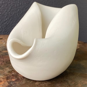 May include: A matte white ceramic vessel with an organic, asymmetrical shape. The object features curved, folded walls that create a deep central opening and a small, rounded spout-like protrusion on the lower front side, with a smooth surface.