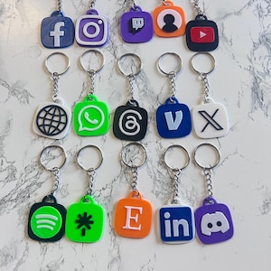 Puede incluir: Una colección de llaveros con varios logotipos de redes sociales, como Facebook, Instagram y YouTube. Cada llavero tiene un anillo y una cadena plateados, con los logotipos en diferentes colores y diseños. Los llaveros están dispuestos sobre una superficie blanca y gris.