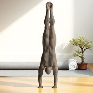 Pode incluir: Escultura de uma pessoa em posição de parada de mãos. A figura é de cor marrom suave. Um tapete de ioga cinza enrolado e um pequeno bonsai em vaso estão no fundo. Uma toalha branca está enrolada perto da árvore.