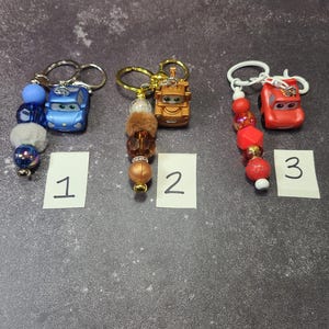May include: Three keychains featuring toy car charms. One is blue with blue and white beads, the second is gold with brown beads, and the third is red with red and white beads. Each keychain has a number on a white label.