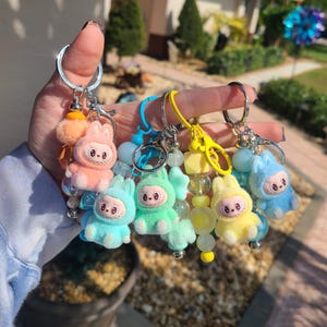 May include: A collection of four keychains featuring fuzzy, pastel-coloured creature charms with wide eyes and toothy grins. Each keychain includes a metal ring, a plastic lobster clasp, and a strand of colourful beads in matching shades of peach, blue, green, and yellow.