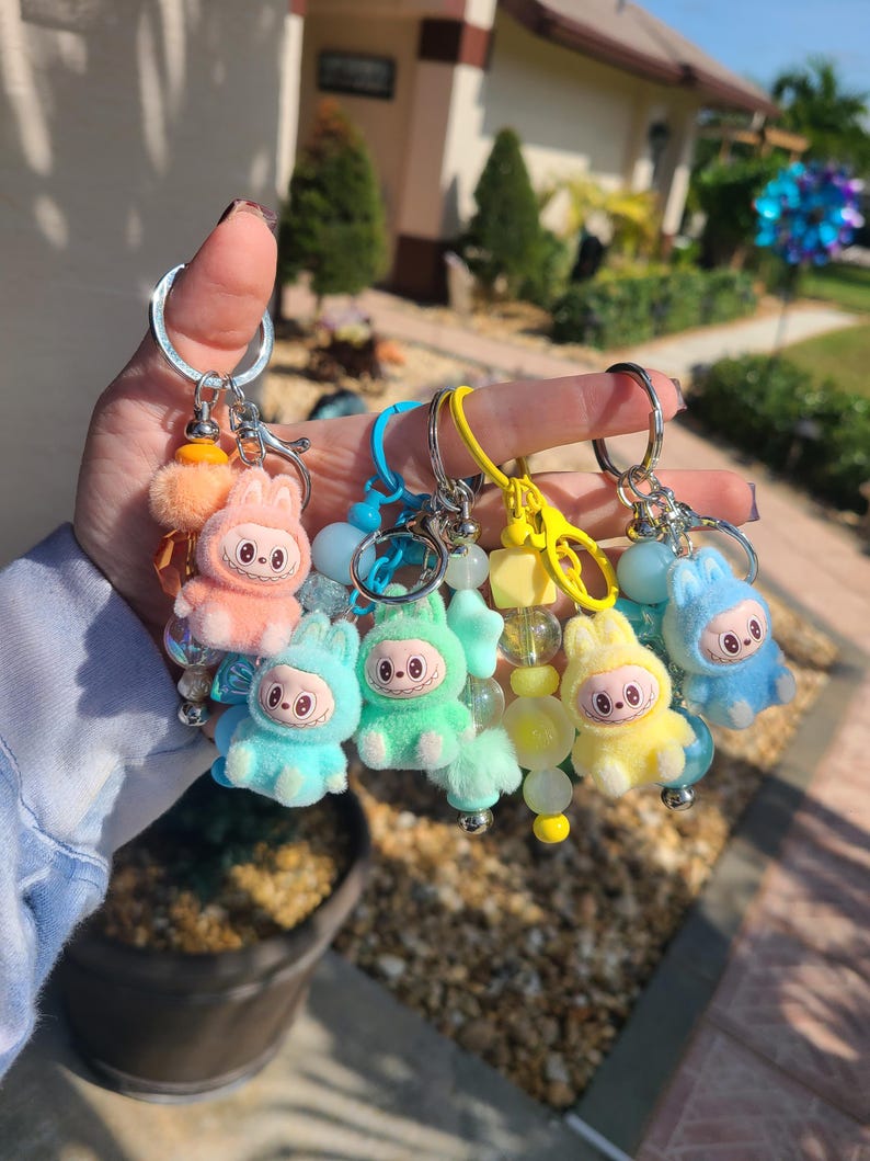May include: A collection of four keychains featuring fuzzy, pastel-coloured creature charms with wide eyes and toothy grins. Each keychain includes a metal ring, a plastic lobster clasp, and a strand of colourful beads in matching shades of peach, blue, green, and yellow.