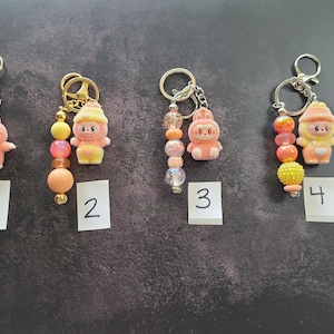 May include: Four keychains featuring small cartoon character charms in pink and yellow hues. Each keychain includes a metal ring and a strand of colourful beads in shades of orange, yellow, and pink. The charms are numbered one through four.