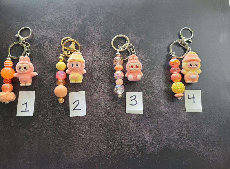 May include: Four keychains featuring small cartoon character charms in pink and yellow hues. Each keychain includes a metal ring and a strand of colourful beads in shades of orange, yellow, and pink. The charms are numbered one through four.