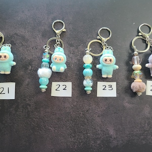 May include: Four metal keychains labelled 21, 22, 23, and 24. Each features a small fuzzy creature figurine in blue or lavender and a strand of decorative beads in various shades of blue, white, and purple with silver-toned metal hardware.