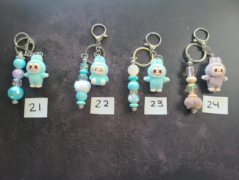May include: Four metal keychains labelled 21, 22, 23, and 24. Each features a small fuzzy creature figurine in blue or lavender and a strand of decorative beads in various shades of blue, white, and purple with silver-toned metal hardware.