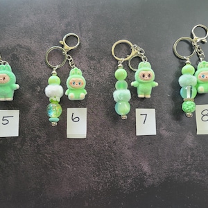 May include: Four keychains labelled 5, 6, 7, and 8, each featuring a light green flocked animal figurine and a strand of decorative beads. The beads vary in texture, including solid, translucent, and fuzzy spheres in shades of green and white, all attached to silver-toned metal key rings.