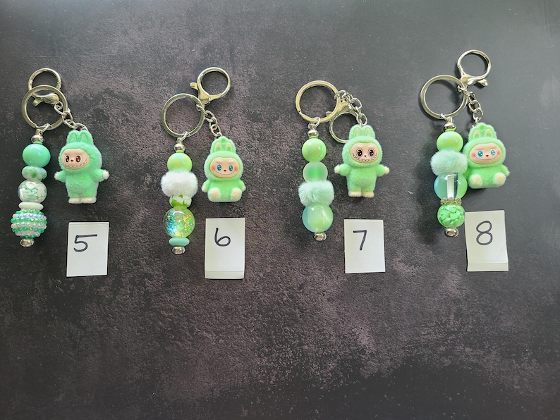 May include: Four keychains labelled 5, 6, 7, and 8, each featuring a light green flocked animal figurine and a strand of decorative beads. The beads vary in texture, including solid, translucent, and fuzzy spheres in shades of green and white, all attached to silver-toned metal key rings.