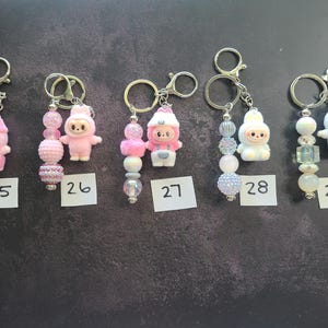 May include: Five keychains labelled 25 through 29, each featuring a small character figurine and a strand of decorative beads. The figurines include pink and white bunny-like creatures. The beads vary in colour, texture, and finish, including clear, iridescent, and solid pink or white spheres attached to silver-toned metal rings.