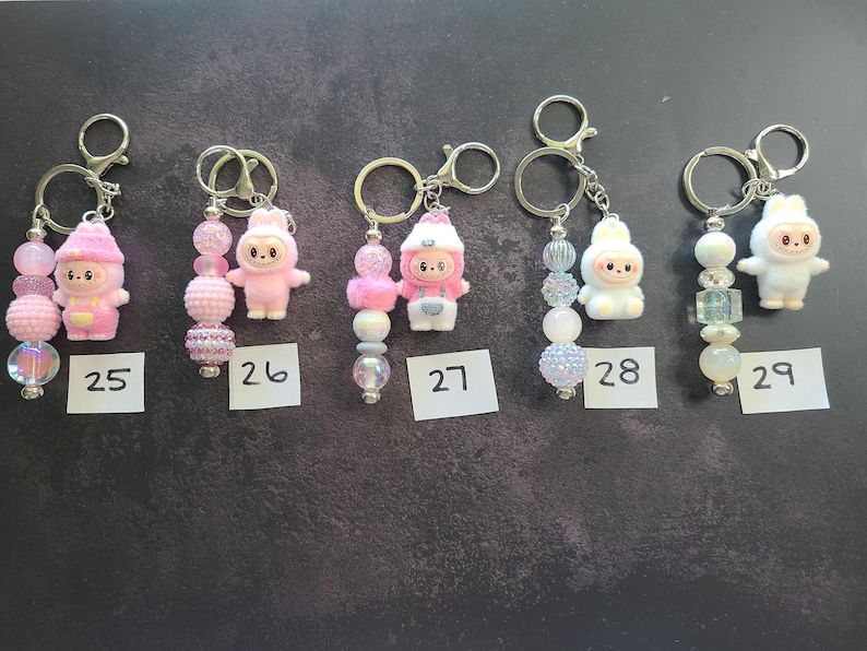 May include: Five keychains labelled 25 through 29, each featuring a small character figurine and a strand of decorative beads. The figurines include pink and white bunny-like creatures. The beads vary in colour, texture, and finish, including clear, iridescent, and solid pink or white spheres attached to silver-toned metal rings.