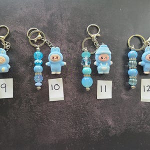 May include: Four keychains labelled 9, 10, 11, and 12, each featuring a small blue character figurine in a hooded outfit attached to a metal ring with a strand of blue beads. The beads vary in texture, including clear, opaque, and glittery finishes.