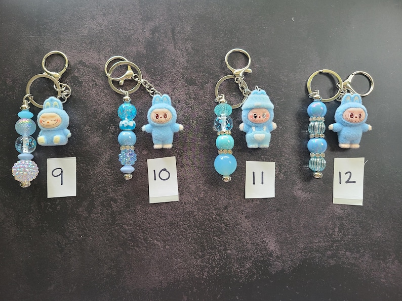 May include: Four keychains labelled 9, 10, 11, and 12, each featuring a small blue character figurine in a hooded outfit attached to a metal ring with a strand of blue beads. The beads vary in texture, including clear, opaque, and glittery finishes.