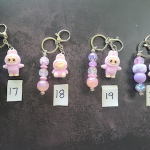 May include: Four keychains featuring light purple bunny-themed figurines and assorted decorative beads in shades of pink, purple, and iridescent white. Each keychain includes a metal ring and is labelled with a small white paper tag numbered 17 to 20.