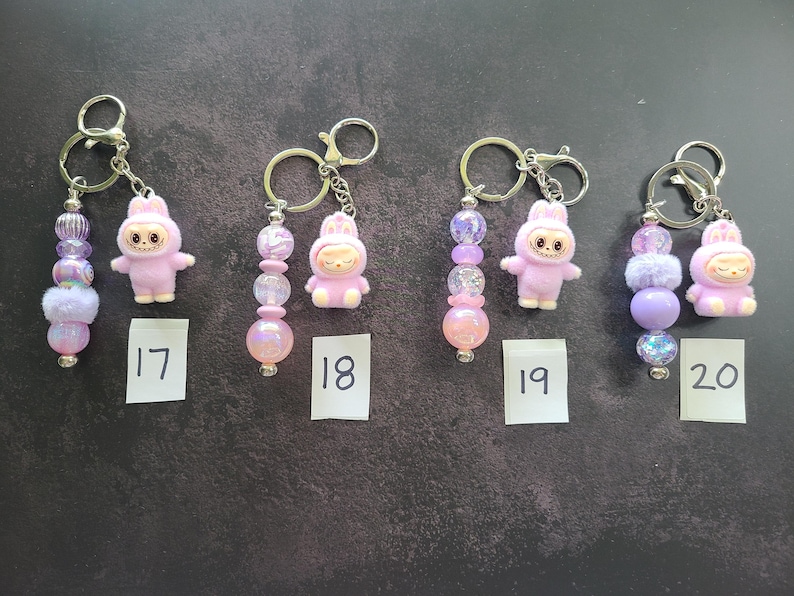 May include: Four keychains featuring light purple bunny-themed figurines and assorted decorative beads in shades of pink, purple, and iridescent white. Each keychain includes a metal ring and is labelled with a small white paper tag numbered 17 to 20.