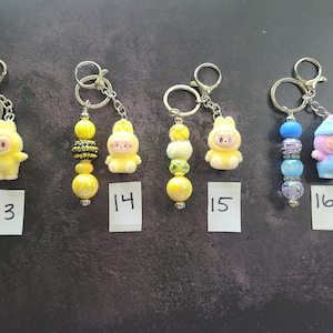 May include: Four keychains labelled 13, 14, 15, and 16. Each features a metal ring, a small character figurine, and a strand of beads. Keychain 13 has yellow and white beads. Keychain 14 has yellow and black beads. Keychain 15 has yellow beads. Keychain 16 has blue beads.