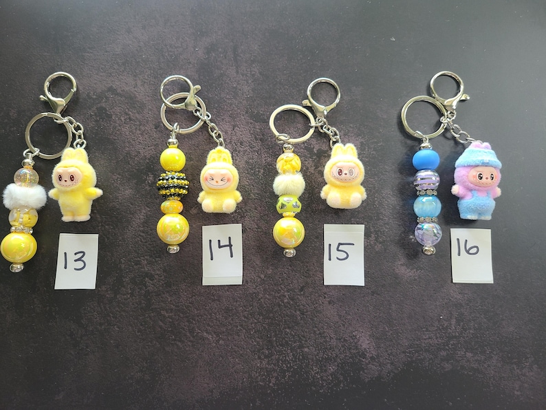 May include: Four keychains labelled 13, 14, 15, and 16. Each features a metal ring, a small character figurine, and a strand of beads. Keychain 13 has yellow and white beads. Keychain 14 has yellow and black beads. Keychain 15 has yellow beads. Keychain 16 has blue beads.