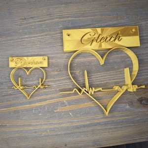 May include: Two gold-coloured heart-shaped metal wall decorations with a heartbeat line. The larger heart has the word "Gleich" on a separate rectangular plaque. The smaller heart has the word "Traumfrau" on a separate rectangular plaque.