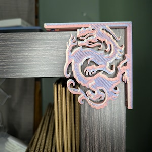 Puede incluir: Un soporte de esquina impreso en 3D con un diseño de dragón. El soporte está hecho de plástico rosa claro y azul y está unido a un estante de madera oscura.