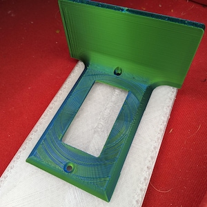 May include: A 3D printed wall plate with a green and blue finish. The plate has a rectangular opening for an electrical outlet.