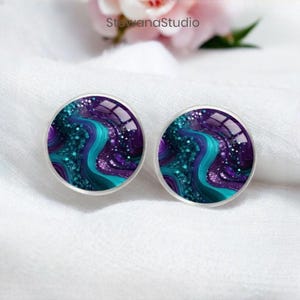 Puede incluir: Un par de pendientes redondos con un diseño abstracto en morado y turquesa. El diseño presenta líneas en espiral y purpurina.