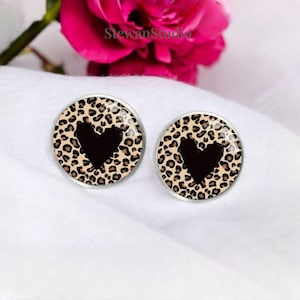 Puede incluir: Un par de pendientes de botón redondos. Cada pendiente presenta un corazón negro en el centro, rodeado de un estampado de leopardo marrón y beige. Los pendientes tienen un borde de metal plateado y están sobre un fondo blanco.