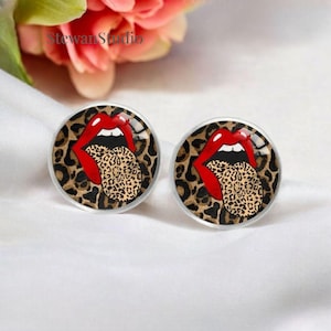 Puede incluir: Un par de pendientes redondos de vidrio con una boca roja abierta y una lengua con estampado de leopardo que sobresale. El fondo de los pendientes es un estampado de leopardo.