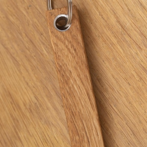 Könnte beinhalten: Ein rechteckiger Holzschlüsselanhänger mit einem silbernen Metallring. Die Holzmaserung ist sichtbar, und der Schlüsselanhänger hat eine hellbraune Farbe. Der Metallring ist mit einer Metallöse am Holz befestigt.