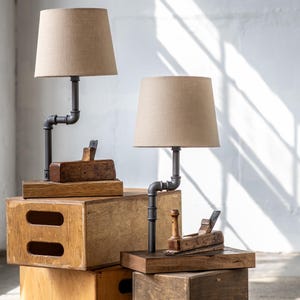 Puede incluir: Dos lámparas de estilo industrial con pantallas de tela beige. Las lámparas presentan un diseño de tubería de metal oscuro y se asientan sobre bases de madera. Cepillos de mano de madera antiguos se colocan cerca de la base de cada lámpara.