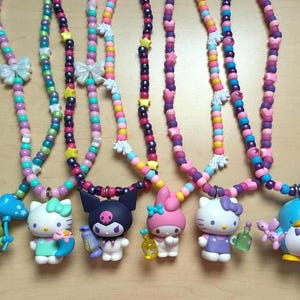 Pode incluir: Colares de contas coloridas com vários pingentes de personagens. Os colares apresentam contas em tons de rosa, roxo, azul e amarelo. Os pingentes incluem um sapo, Hello Kitty, um personagem com uma caveira, My Melody e um pinguim.