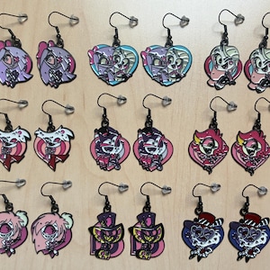 May include: A collection of colorful enamel earrings with black hooks. The earrings feature various cartoon character designs in shades of pink, purple, blue, and red. The earrings are displayed on a light brown surface.