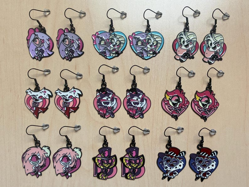 May include: A collection of colorful enamel earrings with black hooks. The earrings feature various cartoon character designs in shades of pink, purple, blue, and red. The earrings are displayed on a light brown surface.