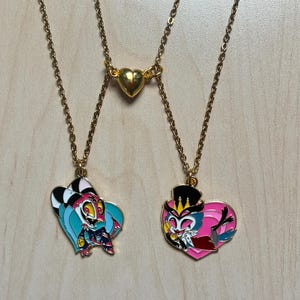 May include: Gold-colored chain necklaces with pendant charms. One necklace features a heart-shaped charm, while the other two have cartoon character pendants. The pendants are colorful with black, pink, blue, and yellow accents.