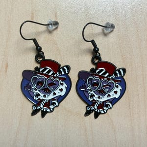 May include: A pair of black dangle earrings. Each earring features a cartoon character with a purple face, heart-shaped glasses, and a red and black top hat. The character is set against a blue heart-shaped background.