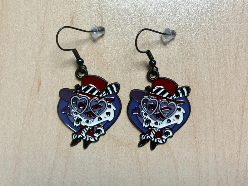 May include: A pair of black dangle earrings. Each earring features a cartoon character with a purple face, heart-shaped glasses, and a red and black top hat. The character is set against a blue heart-shaped background.