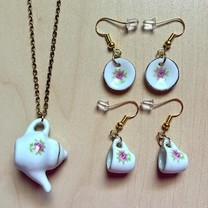 Puede incluir: Un colgante de collar de porcelana blanca con forma de tetera, con una cadena dorada y dos pares de pendientes. Un par son platos pequeños y el otro son tazas pequeñas. Todas las piezas tienen un diseño de rosa rosa.