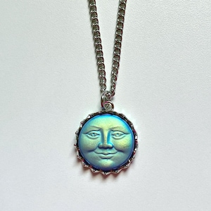 May include: A silver necklace with a pendant depicting a smiling moon. The moon pendant has a blue and green iridescent finish and is set in a decorative silver frame. The chain is a classic link style.